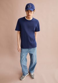 Marineblauw katoenen T-shirt met korte mouwen, gecombineerd met lichtblauwe jeans. Grijze sneakers met witte veters en een marineblauwe pet met borduursel.