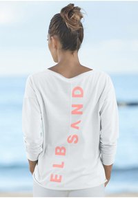 Nainen, jolla on hiukset nutturalla, pukeutuneena valkoiseen pitkähihaiseen paitaan, jossa on "ELBSAND" korallinvärisillä kirjaimilla pystysuoraan takana, seisoo meren äärellä.
