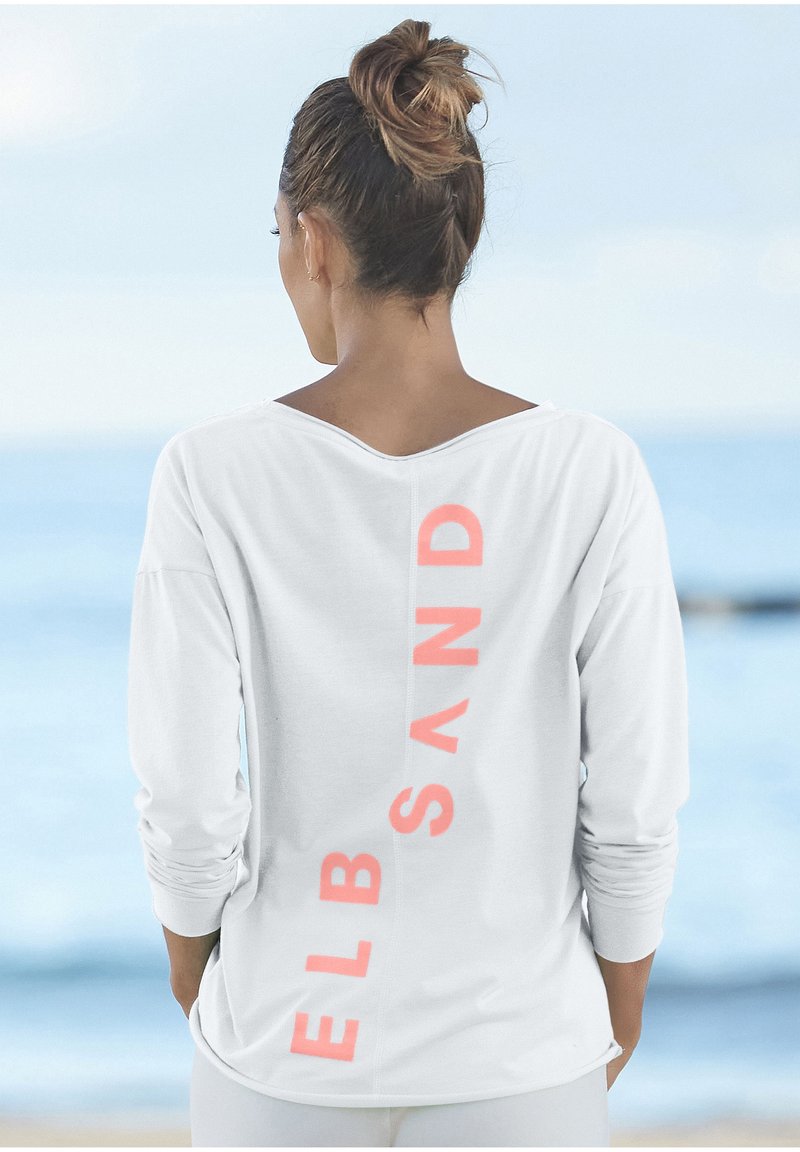 Nainen, jolla on hiukset nutturalla, pukeutuneena valkoiseen pitkähihaiseen paitaan, jossa on "ELBSAND" korallinvärisillä kirjaimilla pystysuoraan takana, seisoo meren äärellä.
