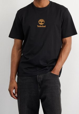 Mężczyzna w czarnej koszulce Timberland z beżowym logo drzewa i napisem, w ciemnych dżinsach, stoi na tle jednolitej jasnej ściany.