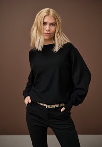 Szőke nő fekete hosszú ujjú felsőben és fekete nadrágban, mintás övvel, zsebében tartott kezével áll barna háttér előtt.