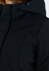 Černá bunda s vysokým límcem a nastavitelnou kapucí. Vyrobena z hladkého, strukturovaného materiálu. Obsahuje švy a minimální kovové detaily.