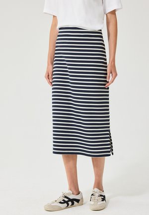 Kvinde iført en navy- og hvidstribet midi-nederdel, hvid top og hvide sneakers med sorte detaljer, stående på en ensfarvet baggrund.