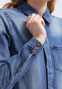 Denimskjorta med spetskrage, knäppte manschetter med bruna detaljer och en bröstficka. Tyget har ett strukturerat, blekt blått utseende.