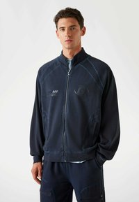 Tumed zip-up jakk pehmeast materjalist, millel on kõrge kaelus, kontrastse õmblusega ja esiküljel taskud koos diskreetsete logo detailidega.