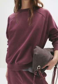 Felpa bordeaux con scollo rotondo e polsini a coste, abbinata a una morbida borsa grigia con chiusura a coulisse e superficie testurizzata.