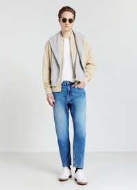 Giovane uomo con occhiali da sole, giacca beige, maglione grigio sulle spalle, camicia bianca, jeans blu e sneakers bianche, in piedi con una mano in tasca.