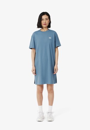 Robe bleue à manches courtes en coton doux, avec un col rond, des fentes sur les côtés et un petit logo sur la poitrine. Portée avec des baskets blanches.