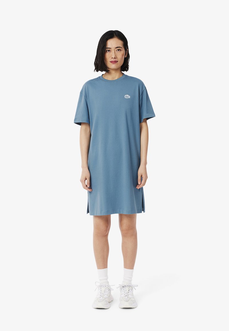 Abito blu a maniche corte in morbido cotone, con scollatura a girocollo, fessure laterali e un piccolo logo sul petto. Indossato con sneakers bianche.