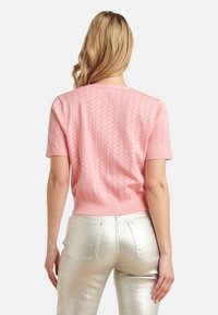 Rosa kurzärmeliger Pullover mit strukturierten Mustern und geripptem Saum, kombiniert mit glänzenden, silbernen Hosen, von hinten betrachtet.