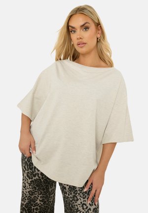 Lys beige oversized t-shirt med bred halsudskæring og korte ærmer, lavet af blødt, tekstureret stof, båret med mønstrede sorte og grå bukser.