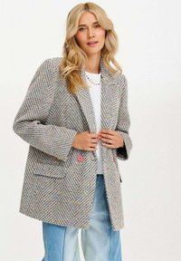 Blondýnka v texturovaném, vícebarevném tvídovém saku s růžovými knoflíky, které má přes bílé tričko a světle modré džíny, na bílém pozadí.