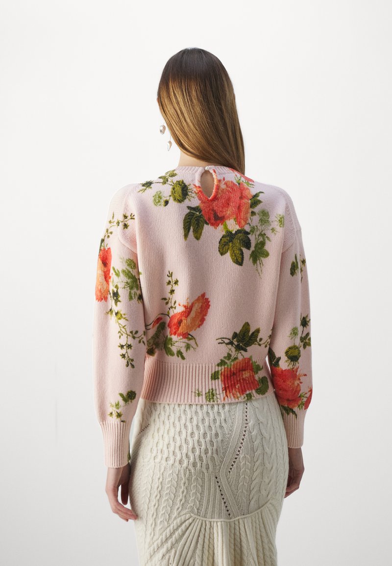 Pull en tricot rose avec des motifs floraux, comprenant des roses rouges et des feuilles vertes. Manches et ourlet côtelés, avec un détail en forme de trou à l'arrière.