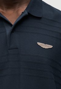 Μπλε ναυτικό πόλο με υφασμάτινη υφή, που διαθέτει οριζόντιες ρίγες και ένα μπεζ σήμα με το λογότυπο της Aston Martin στο αριστερό μέρος του στήθους.