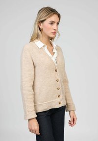 Cardigan en laine beige avec un décolleté en V profond, des poignets et un ourlet côtelés ; il comporte des boutons marron et est porté avec une chemise blanche à col en dessous.