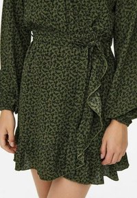 Robe portefeuille verte à imprimé floral, confectionnée dans un tissu léger. Elle présente des manches longues, un ourlet volanté et une taille nouée avec un motif floral subtil.