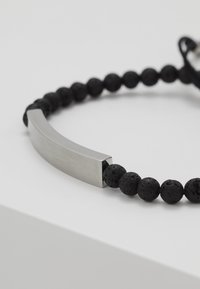 Pulsera de cuentas de piedra de lava negra con un elemento metálico curvado en el centro y un cierre de cordón ajustable. Las cuentas texturizadas contrastan con el metal suave.