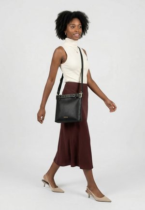 Kvinde går iført en ærmeløs cremefarvet rullekrave, brun midi-nederdel, beige hæle og bærer en sort crossbody taske i læder med spændedetalje.