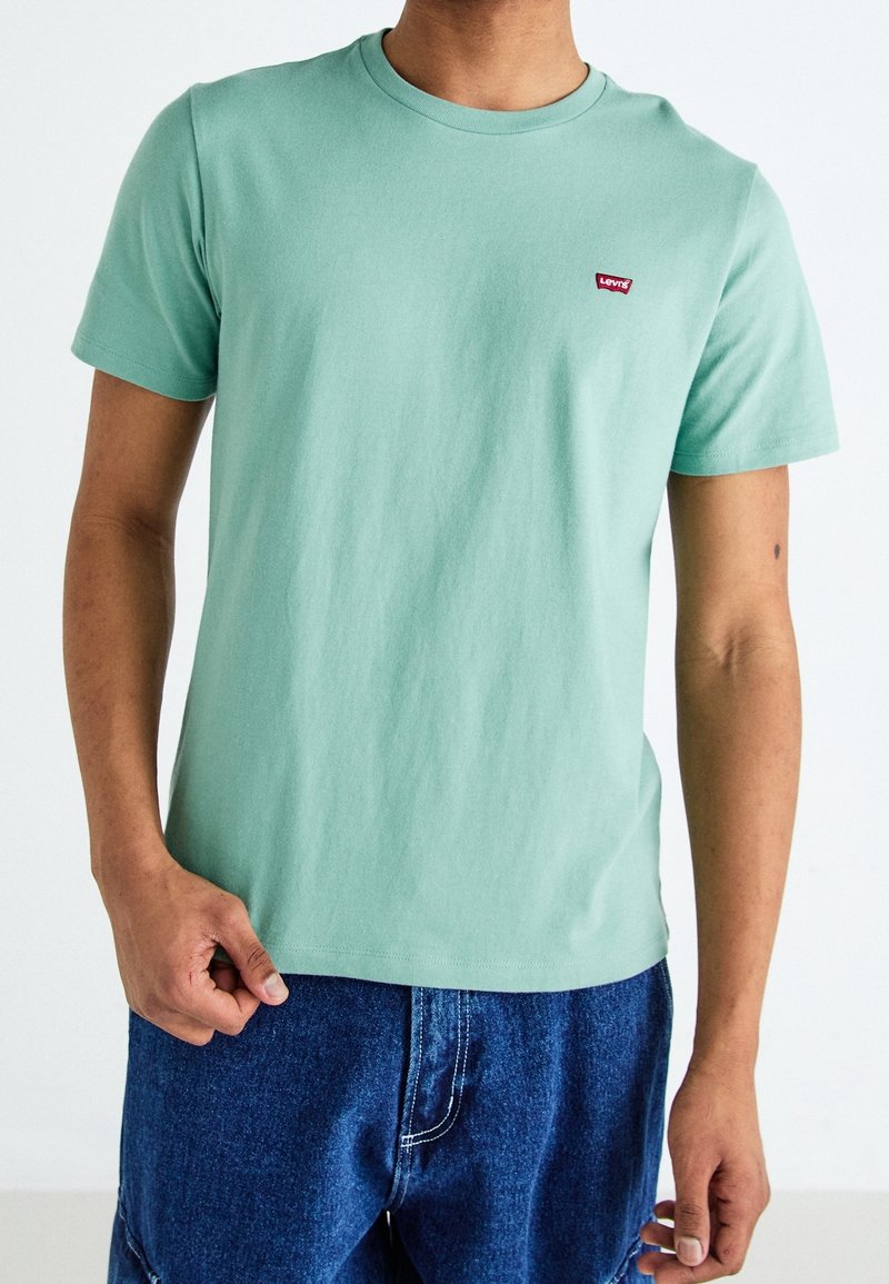 Personne portant un t-shirt Levi's vert clair à manches courtes et un jean bleu en denim, debout devant un fond blanc uni.