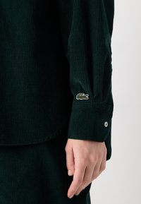 Manica di camicia in corduroy verde scuro con un polsino con bottone argento e un piccolo logo di coccodrillo ricamato in bianco e verde.