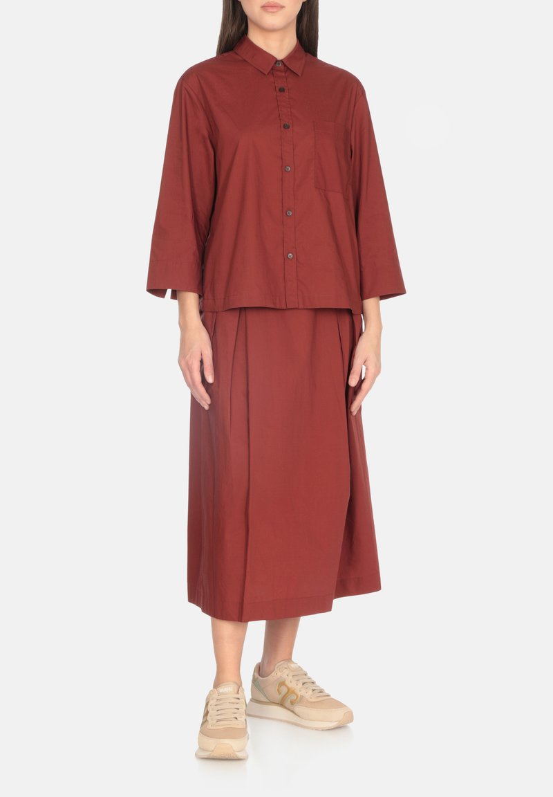 Camicia di cotone bordeaux con colletto, chiusura con bottoni e maniche a tre quarti, abbinata a una lunga gonna coordinata e sneakers beige.