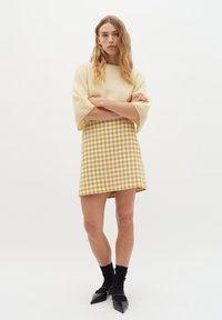 Nertinis kremas megztinis su plačiomis rankovėmis, derinamas su geltonu ir baltu houndstooth raštuotu sijonu. Modelis dėvi juodas kulkšnines kojines ir smailias flats avalynes.