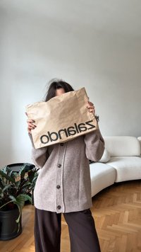 Pessoa com um cardigan bege e calças escuras a segurar um saco de papel castanho amarrotado com o texto "Zalando", a estar em uma sala de estar minimalista.