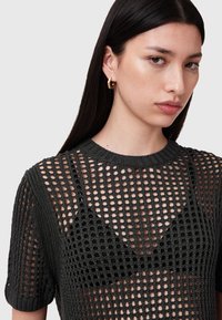 Sort strikket top med et bredt mesh-mønster, korte ærmer og en rund halsudskæring, lagd over en sort bralette. Hud synlig gennem vævningen.