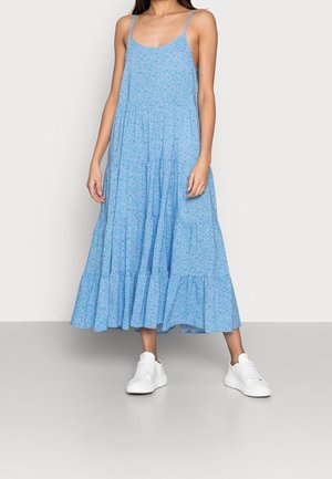 Femme portant une robe midi sans manches à fleurs bleu clair avec bretelles spaghetti et baskets blanches, debout sur un sol gris.