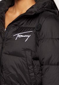Czarna kurtka puchowa z kapturem, wyposażona w zamek błyskawiczny, regulowany sznurek oraz biały haftowany logo "Tommy" na piersi.