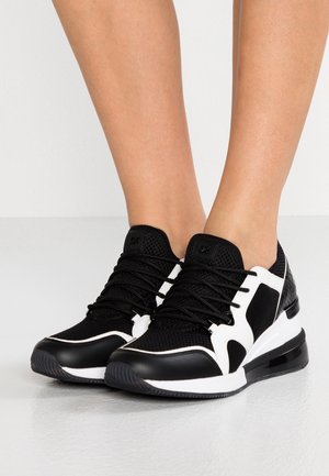Schwarz-weiße sportliche Schnürschuhe, getragen auf nackten Füßen vor einem schlichten hellen Hintergrund.