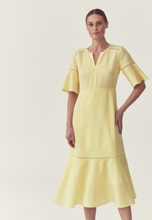 Nő halványsárga midi ruhában, bővülő ujjakkal és szegéllyel, finom kivágott részletekkel az ujjakon és a ruha aljánál.