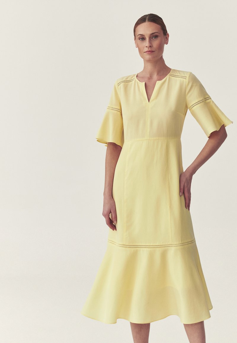 Femme portant une robe midi jaune pâle avec des manches évasées et un ourlet amply, comportant de délicats détails en découpe sur les manches et près du bas.