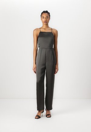 Tamnozeleni saten jumpsuit s tankim naramenicama, prilagođenim tijelom i širokim nogavicama. Ima blagi sjaj i bočne šavne džepove. Crne sandale s potpeticama.