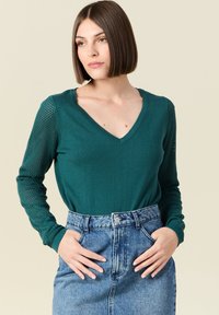 Maglietta lunga teal a maniche con scollo a V e maniche in maglia testurizzata abbinata a una gonna di jeans a vita alta con chiusura a bottoni.
