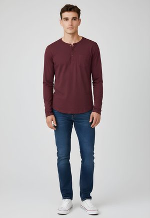 Joven de pie, vestido con una camiseta henley de manga larga color granate, vaqueros azules y zapatillas blancas sobre un fondo claro y sencillo.