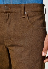 Pantalones de denim marrón con textura, de cintura alta, con bolsillos frontales y cierre de botón metálico. Presentan costuras en contraste y un ajuste estructurado.