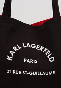 Czarna torba typu tote z czerwonym wnętrzem, z białym napisem: "KARL LAGERFELD PARIS 21 RUE ST-GUILLAUME" w zakrzywionym ułożeniu.