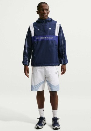 Mand stående iført en mørkeblå og hvid Nike Shox jakke, hvide og lyseblå shorts, hvide Nike sokker og blå og hvide atletiske sko.