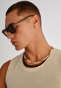 Νεαρός άνδρας που φοράει μαύρα γυαλιά ηλίου, κολιέ με χάντρες και αμάνικη μπεζ μπλούζα με σκούρο μπλουζάκι από κάτω, μπροστά σε απλή φόντο.