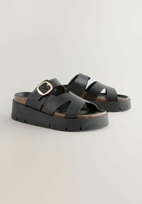 Sandali platform neri con design a strappi incrociati, dotati di un accento in fibbia dorata e plantare in sughero. Suola in gomma testurizzata per una buona aderenza.