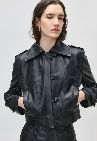 Mladić s valovitom smeđom kosom, odjeven u crnu kožnu jaknu s gumbima, ruke u džepovima i srebrne naušnice u obliku naušnica.