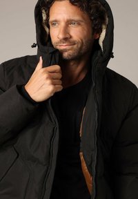 Veste matelassée noire avec fermeture éclair à l'avant, dotée d'une doublure orange à motif camo et d'une capuche douillette en fourrure. Texture lisse, matériau durable.