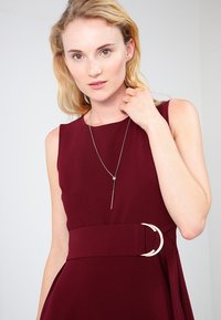 Burgundy dress with a fitted bodice and flared skirt, featuring a wide waistband with a circular buckle. Slim silver necklace with a pendant.