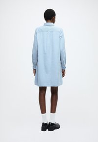 Rochie albastru deschis cu mâneci lungi, țesătură texturată, manșete cu nasturi și guler; siluetă dreaptă care se oprește deasupra genunchiului.