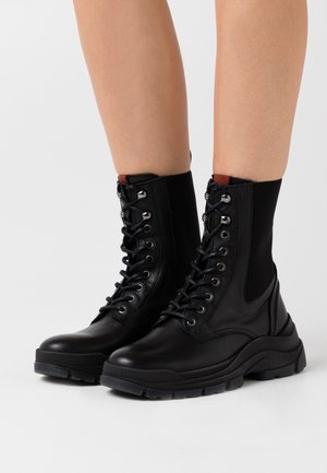 Black ankle boots with a textured leather upper, mesh accents, lace-up design, and thick rubber outsole. Silver eyelets and red heel tab detail.