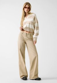 Pull court beige à rayures avec col en V, associé à un pantalon large marron clair. Le mannequin porte des lunettes de soleil foncées et des chaussures noires.