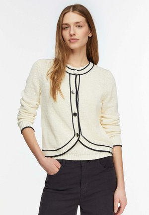 Jeune femme aux longs cheveux portant un cardigan crème avec bordures et boutons noirs, associé à un pantalon noir, debout avec une main dans la poche.