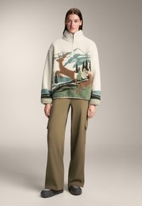 Bluza polarowa z wysokim kołnierzem w kolorze kremowym, z nadrukiem krajobrazu górskiego i jeleni w zielonym i brązowym kolorze. W zestawie z oliwkowymi spodniami cargo.