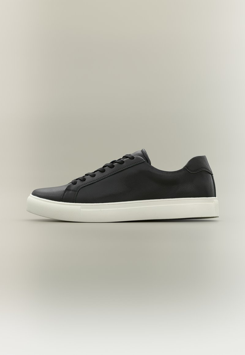 Baskets basses noires avec semelle en caoutchouc blanche et lacets noirs, présentées de profil sur un fond neutre.